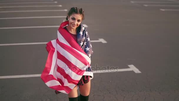 Гордая американская девушка ходит и заворачивает американский флаг через плечи, глядя в камеру. Слоумоушн — стоковое видео