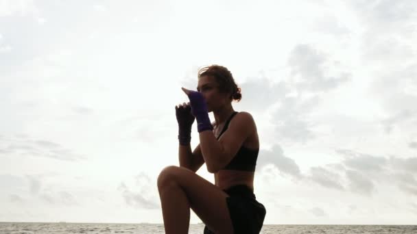 Duro boxeo de sombras de mujer bonita. Hermosa boxeadora entrenando junto al mar en la playa por la mañana, lanzando golpes en cámara lenta — Vídeos de Stock