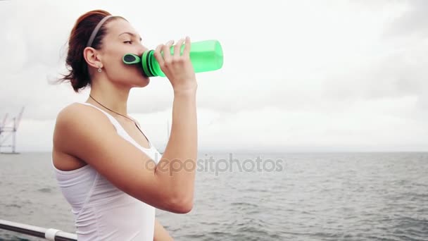 Žíznivý fitness žena odpočívá pauzu s vodou láhev pití po tréninku. Krásná žena školení u moře — Stock video