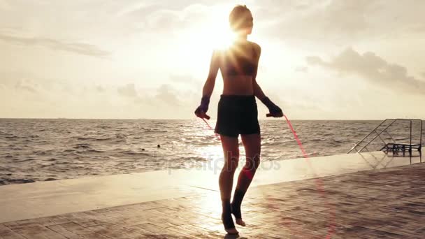 Молодая женщина работает на скакалке против солнца на пляже в медленном темпе. Девушка прыгает на скакалке у моря — стоковое видео