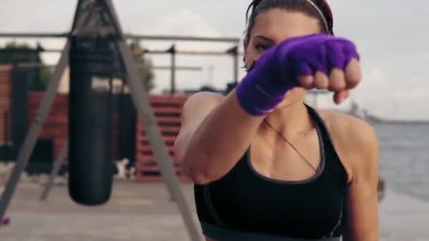 Closeup view of a young woman training with her hands wrapped in purple boxing tapes and looking in the camera. Training by the beach in summer — Stock Video