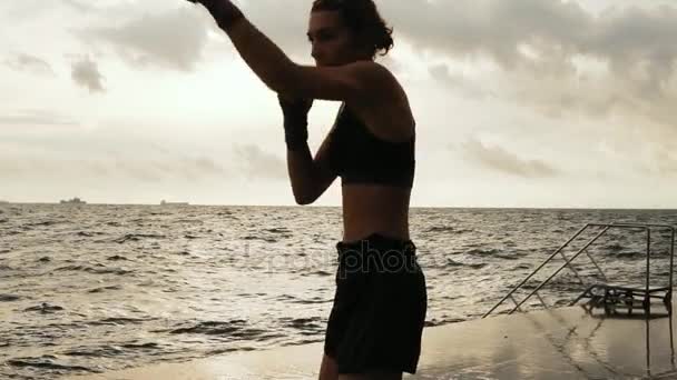 Жінка-боксер робить бічний удар її ніг проти сонця біля моря в повільному русі. Красиві тренування боксерів на пляжі вранці, кидаючи удари — стокове відео
