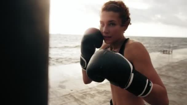 Άποψη closeup της ένα ισχυρό αθλητικό θηλυκό μπόξερ στο γάντια άσκηση με μια τσάντα ενάντια στον ήλιο δίπλα στη θάλασσα. Θηλυκό μπόξερ εκπαίδευση — Αρχείο Βίντεο