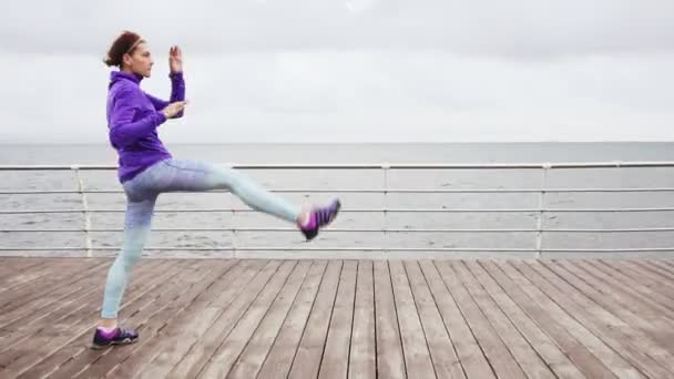 Vídeo de alto ritmo: Mujer joven haciendo ejercicio, pateando piernas y estirándose en la playa por el océano temprano en la mañana — Vídeos de Stock