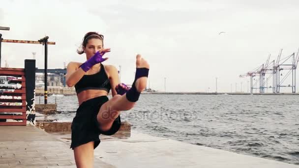 Joven boxeadora practicando patada lateral de sus piernas junto al mar en cámara lenta. Hermosa mujer boxeadora entrenando en la playa por la mañana — Vídeos de Stock