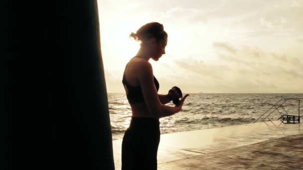 Mulher atlética recebendo seus punhos prontos para as luvas de boxe, envolvendo bandagem em torno deles de pé contra o sol. Boxer feminino envolvendo mãos com invólucros de boxe na praia. Inflação da lente — Vídeo de Stock