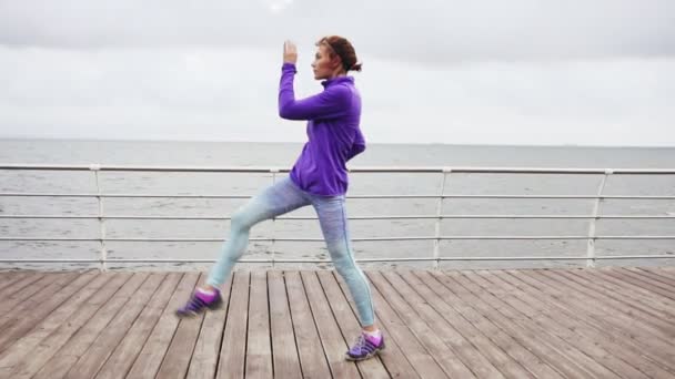 Νεαρή γυναίκα άσκηση, κλωτσώντας τα πόδια και το τέντωμα στην παραλία από τον ωκεανό, νωρίς το πρωί. Slowmotion βολή — Αρχείο Βίντεο