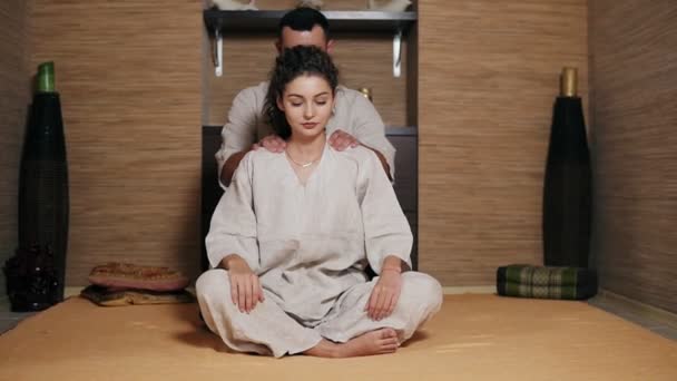 Unga manliga thailändska massagist massera unga kvinnans tillbaka och axlar sitter bakom henne. Vacker flicka tar emot thailändsk massage — Stockvideo
