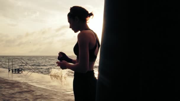 Boxeadora mujer preparando sus puños para los guantes de boxeo envolviendo vendaje alrededor de ellos de pie contra el sol. Mujer joven envolviendo las manos con envolturas de boxeo en la playa. Lente llamarada — Vídeos de Stock