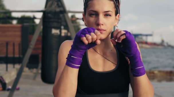 Tiro em câmera lenta: Vista de perto de uma jovem mulher de boxe sombra com as mãos embrulhadas em fitas de boxe olhando para a câmera. Bonito boxeador feminino treinando na praia pela manhã — Vídeo de Stock