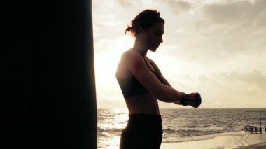 Kamera hareketi yuvarlak: kadın boksör boks ile el sarma güneş karşı okyanus durarak sahilde tamamladı. Mercek parlaması