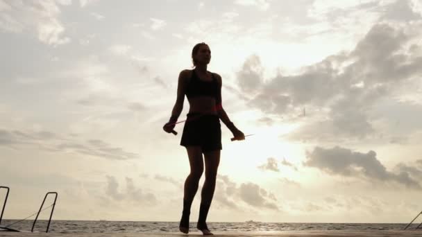 Mujer atlética irreconocible haciendo ejercicio en la cuerda de salto contra el sol en la playa en cámara lenta. Chica saltando en una cuerda de saltar por el mar — Vídeos de Stock