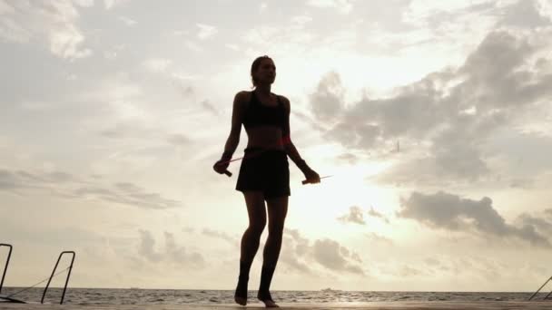 Ugjenkjennelig atletisk kvinne som trener på hoppetauet mot solen ved stranden i sakte bevegelse. Jente hopper på et hoppetau ved sjøen – stockvideo