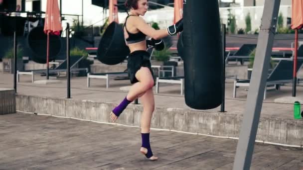 Hoog tempo video: sterke atletische vrouwelijke bokser in handschoenen uitoefenend met een zak. Ze raakt een zak met haar been. Training buiten. Vrouwelijke bokser die traint. Tikje schot — Stockvideo