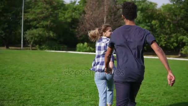 Vista posterior siguiente disparo de linda pareja internacional atractiva cogidas de la mano y corriendo en el campo con hierba verde en el parque — Vídeo de stock