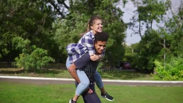 Bonito afro cara piggybacking sua namorada feliz caucasiano e girando em torno de correr na grama verde no parque ensolarado. Data romântica de um feliz casal multiétnico — Vídeo de Stock