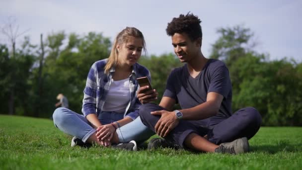 Pareja de carreras mixtas sentados en el césped y selecciona imágenes en su teléfono inteligente. Los jóvenes sonríen y ríen. Lindos amigos descansando en un parque, sentados en la hierba, disfrutando de un teléfono inteligente — Vídeo de stock