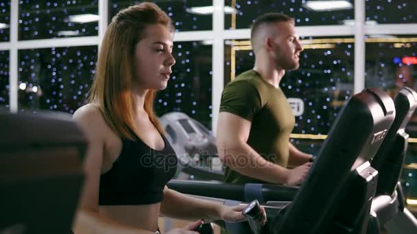 Attractive caucasian girl is starting to run on the treadmill in the sport gym. Young attractive man is running behind. Slowmotion shot — Stock Video