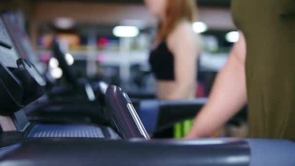 Primer plano de una cinta de correr con un hombre irreconocible caminando sobre ella en el gimnasio deportivo calentándose antes del entrenamiento. Joven mujer atractiva está caminando detrás. Disparo en cámara lenta — Vídeo de stock