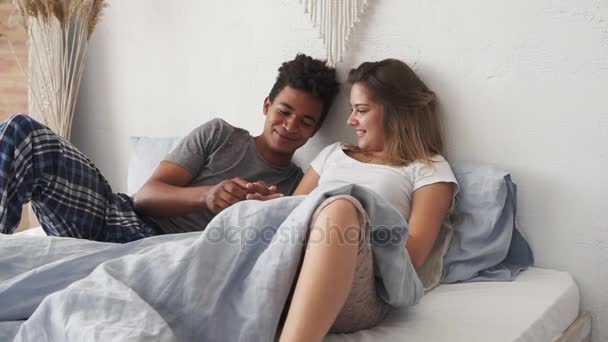 Unga söta multietniskt par i sängen och pratar. Stilig afrikanska man håller sin kaukasiska flickvänner hand och berätta för henne något. Härlig man och kvinna tittar på varandra med kärlek — Stockvideo