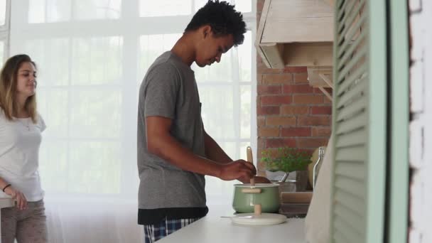 Africký muž připravuje jídlo v hrnci, stojící v kuchyni. Jeho kavkazské přítelkyně přijde k němu a objímat. Skutečná láska. Slowmotion shot — Stock video