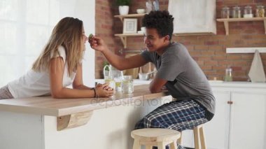 Genç çok ırklı çift mutfak masasının sabah konuşuyor. Yakışıklı adam eğleniyor kız alnındaki yeşil yaprak. Slowmotion atış