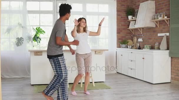 Счастливая мультирасовая пара танцует на кухне в пижаме, слушает музыку по утрам дома. Слоумоушн — стоковое видео