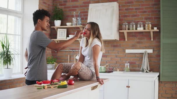 Slowmotion shot of attractive multiethnic couple chatting in the kitchen early in the morning. Handsome man feeding his girlfriend while cooking breakfast — Stock Video
