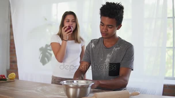 Homem africano atraente preparando massa na mesa de madeira. A namorada está a provocá-lo com a maçã. Jovem casal multiétnico se divertindo. Tiro em câmara lenta — Vídeo de Stock