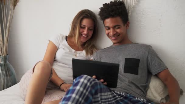 Joven pareja multiétnica riendo viendo películas en la cama en una tableta digital. Disparo en cámara lenta — Vídeos de Stock