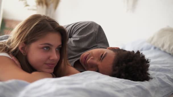 Vista de cerca de la feliz pareja multirracial acostada en la cama y hablando. Atractivo joven hombre y mujer enamorados. Concepto romántico — Vídeo de stock