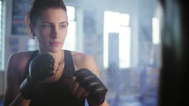 Vista da vicino del pugile caucasico che colpisce il sacco da boxe con le mani nei guanti in palestra con il fumo. Duro allenamento di potenza di un kickboxer femminile — Video Stock