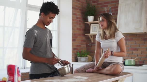 Jovem casal multiétnico cozinhar juntos na cozinha. Homem africano atraente misturando ingredientes em uma panela. A namorada dele está sentada na mesa. Estão a falar e a divertir-se. Tiro em câmara lenta — Vídeo de Stock