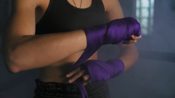 Vista da vicino delle mani femminili che vengono avvolte per la boxe in una stanza buia con fumo. Giovane donna in forma che avvolge le mani con benda da boxe viola. Girato in 4k — Video Stock