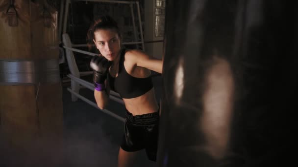 Медленный снимок женщины-боксера в перчатках, наносящей удар кулаком по боксерской сумке во время тренировки в темной фитнес-студии с дымом — стоковое видео