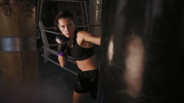 Slowmotion skjuten av en kvinnlig boxare i handskar hiting hårt en boxning väska med hennes knytnäve medan utbildning i en mörk fitnesstudio med rök — Stockvideo