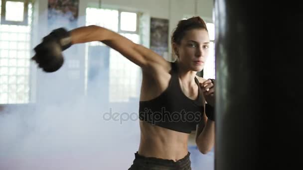Jovem boxeadora caucasiana batendo no saco de boxe com as mãos em luvas no ginásio com fumaça. Treinamento de poder difícil de um kickboxer feminino — Vídeo de Stock