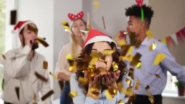 Молодая привлекательная женщина выдувает золотые конфетти из рук в рождественской шляпе, в то время как ее коллеги тусуются на заднем плане в офисе, празднуя Рождество и Новый год. Слоумоушн — стоковое видео