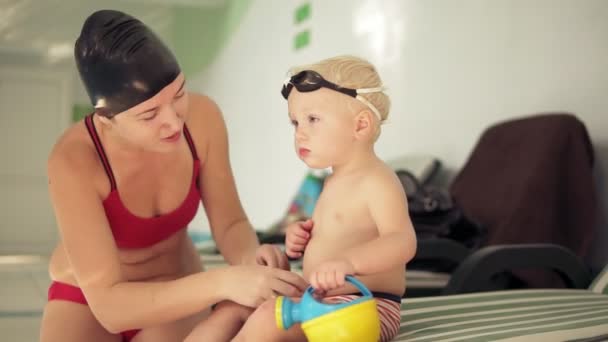 Młoda matka i jej cute little blond dziecko siedząc przy basenie i przygotowanie do lekcji pływania. Szczęśliwa mama rozmawia z dzieckiem. — Wideo stockowe