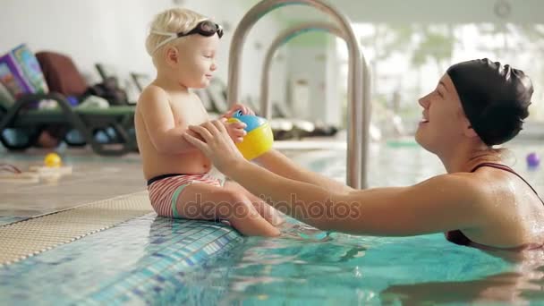 Jovem mãe está ensinando sua linda criança loira a nadar na piscina. Ela está sentada junto à água, então ela o ajuda a mergulhar. Criança bonito está vestindo óculos de proteção especiais na piscina — Vídeo de Stock