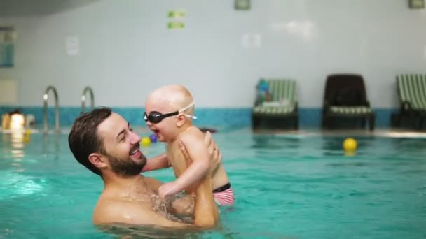 Jovem pai levantando seu menino em óculos de proteção da água enquanto o ensina a nadar na piscina. Menino feliz e seu pai estão rindo e se divertindo — Vídeo de Stock