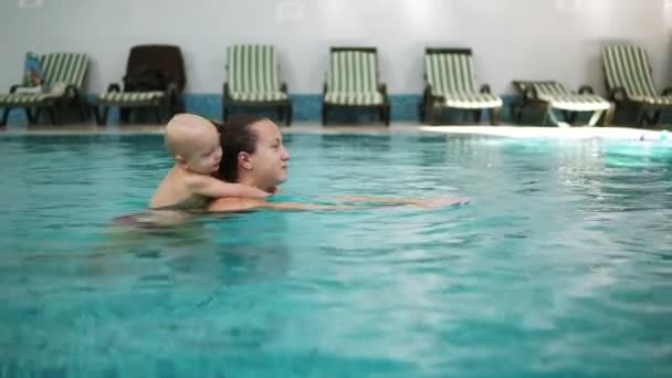 La joven madre está nadando en la piscina sosteniendo a su pequeño y lindo niño en su espalda. Adorable niño pequeño está abrazando a su madre y sosteniéndola mientras nada en la piscina. Cabalgata en el agua — Vídeos de Stock