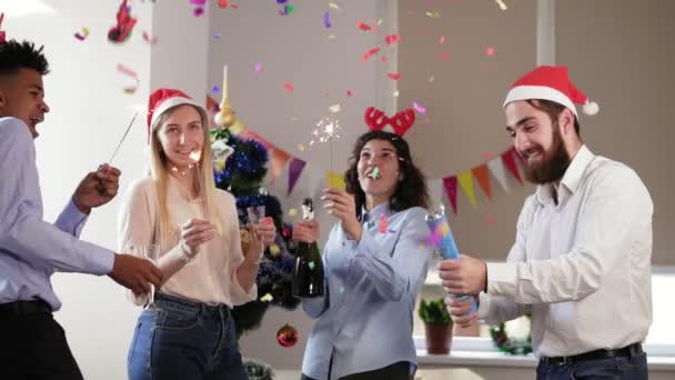 Счастливые мультиэтнические люди в смешных шляпах зажигают искры, празднующие Рождество и Новый год в офисе, взволнованные разнообразные молодые друзья, держащие бокалы для шампанского, стреляющие бенгальскими огнями — стоковое видео