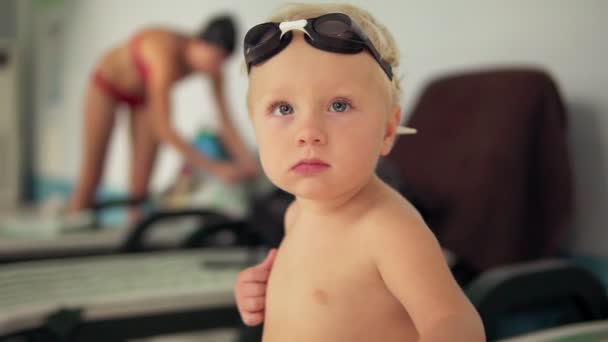 Retrato de un hermoso niño pequeño con gafas sentadas junto a la piscina y mirando a la cámara esperando a que venga su madre. Disparo en cámara lenta — Vídeo de stock