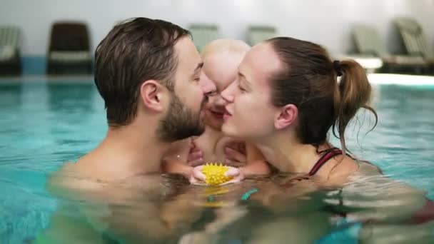 Jovem mãe e pai estão segurando seu filho bonito na piscina e beijando uns aos outros em seus lábios. Criança feliz está sorrindo — Vídeo de Stock