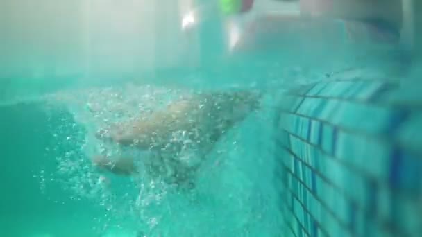 Speelse peuter zit op de rand van het zwembad, de spatten tijdens het schoppen zijn benen in het water. Een onderwater schot — Stockvideo