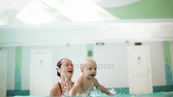 Молодая мать поднимает сына из воды, держа его за ноги, и учит его плавать в бассейне. Счастливый маленький мальчик и его мать смеются и веселятся — стоковое видео
