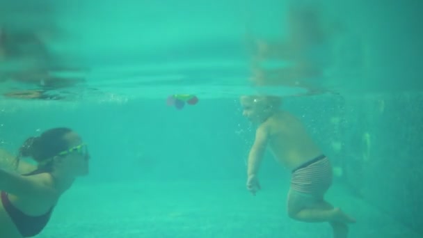 Um tiro subaquático de criança loira bonito nadando sob a água, juntamente com sua mãe na piscina. Então a mãe dele está a tirá-lo da água. A mãe está a ensiná-lo a nadar. — Vídeo de Stock