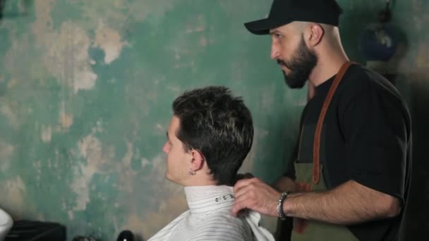 Jovem atraente sentado em uma cadeira enquanto barbeiro barbudo está preparando-o para um corte de cabelo. Barbearia elegante — Vídeo de Stock