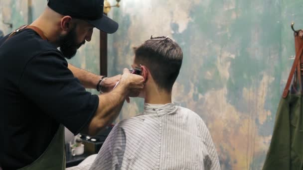 Бородатий перукар створює пряму лінію, використовуючи електричний тример на храмі чоловіка. Молодий красивий кавказький чоловік отримує стрижку в сучасному перукарському магазині. Постріл уповільнення — стокове відео